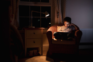 Child reading in his room in the dark while using a lamp.